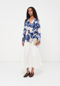 Langärmelige Bluse in Blau und Weiß mit Muster, kombiniert mit einem weißen knielangen Rock und braunen Loafers. Das Model hält eine geflochtene Clutch.