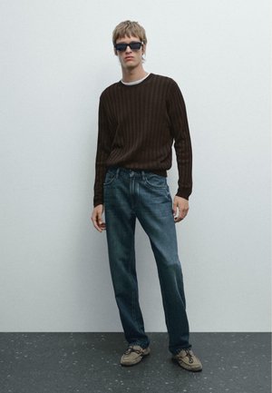 Maglione marrone a costine, jeans blu e scarpe grigie. L'outfit presenta una vestibilità comoda con cuciture a vista e dettagli delle tasche.