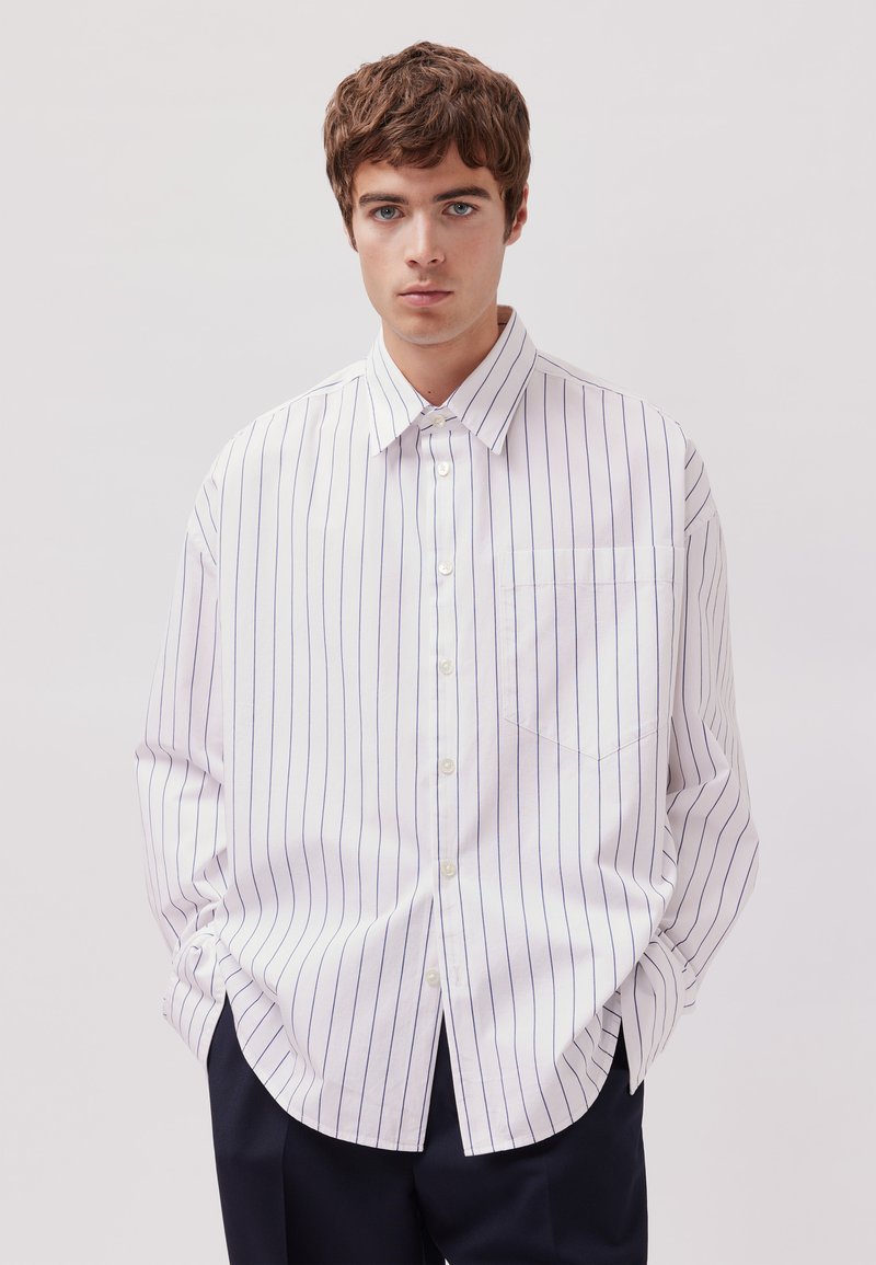 Camicia bianca con righe verticali blu, maniche lunghe, taschino sul petto e orlo curvo. Realizzata in un tessuto liscio.