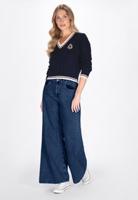 Marineblauwe kabelgebreide sweater met V-hals en witte/rode strepen, gecombineerd met wijde donkere spijkerbroek en witte sneakers. Logo detail op de sweater.
