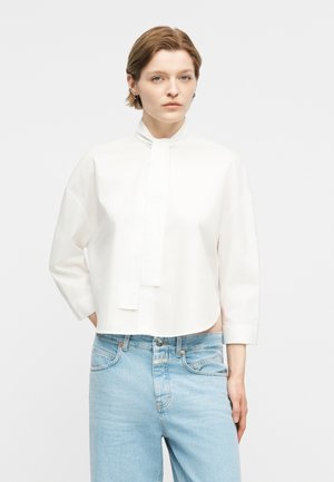 Mladá osoba s krátkymi vlasmi oblečená v bielej blúzke s dlhým rukávom a detailom na krku v podobe kravaty a svetlomodrých džínsach s vysokým pásom, stojaca pred bielym pozadím.