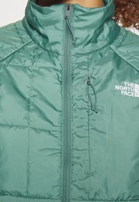 Zelená izolovaná bunda s vysokým límcem, předním zipem a elastickými detaily. Má prošívanou strukturu a logo na hrudi.