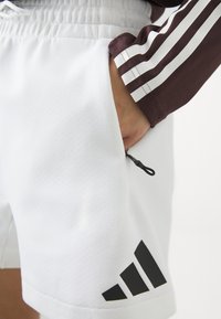 Baltos sportinės šortai su elastinga juosta, šonine kišene ir juodu geometriniu logotipu dešiniajame apatiniame kampe. Lygus paviršius.