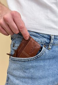 Brauner Leder-Kartenhalter mit Reißverschluss, in einer vorderen Tasche von blauen Jeans platziert. Strukturierte Oberfläche mit sichtbaren Stitching-Details.