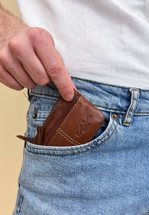 Ruskea nahkainen korttiteline vetoketjulla, sijoitettuna sinisten farkkujen etutaskuun. Pintarakenteen yksityiskohdat ja ompelut näkyvät.