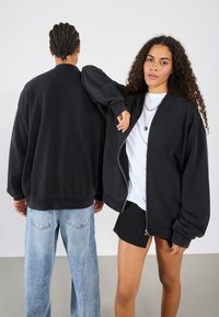 Black oversized zip-up jacket with dropped shoulders, ribbed cuffs, and hem. Paired with light-wash jeans and black shorts. Smooth fabric texture.