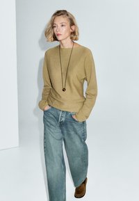 Junge Frau mit kurzen blonden Haaren, die einen beigen Pullover, weite blaue Jeans, braune Schuhe und eine lange Halskette trägt. Sie steht vor einer weißen Wand.