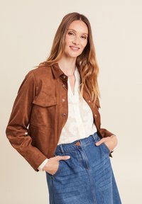 Veste en daim marron avec un col, fermeture à pression à l'avant et deux poches poitrine, portée sur un chemisier blanc et une jupe en denim bleue.