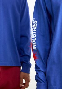 Blaues Baumwollhemd mit langen Ärmeln und grafischem Druck. Mit dem weißen Logo "INDUSTRIES" entlang des Ärmels und einem roten Sonneneffekt gestaltet.