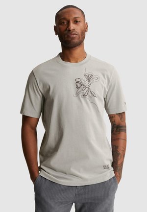 Mand med tatoveringer iført lysegrå T-shirt med abstrakt blomsterdesign på brystet og teksten "Cast Iron" nær kanten, står mod en ensfarvet baggrund.