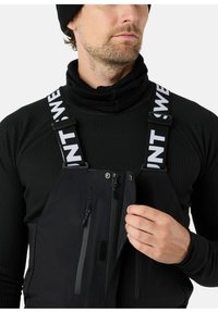 Zwarte geïsoleerde overall met verstelbare banden, voorzien van een wit logo, meerdere met rits afgesloten zakken en een zachte binnenvoering.