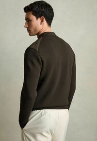 Pulover tricotat maro cu un corp texturat și accente netede pe umeri. Design ajustat cu tiv cu nervuri, purtat cu pantaloni de culoare deschisă.