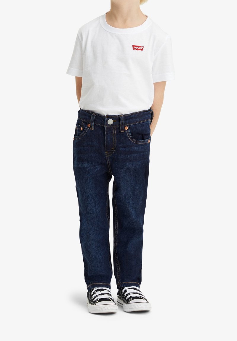 Enfant portant un t-shirt blanc Levi's, un jean bleu foncé et des baskets noires et blanches, debout les mains dans le dos.