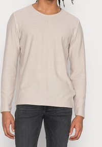 Beiger Pullover mit langen Ärmeln und strukturiertem Muster, Rundhalsausschnitt und kontrastierenden Seitennähten, kombiniert mit dunklen Jeans.