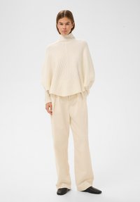 Kobieta stoi ubrana w kremowy sweter z wysokim kołnierzem z prążkowanej tkaniny o szerokich rękawach, beżowe spodnie o szerokich nogawkach oraz czarne buty na kwadratowym nosku na białym tle.