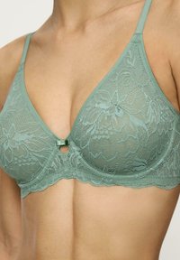 Bralette in pizzo verde morbido con motivo floreale, coppe triangolari, bordi ondulati e un piccolo fiocchetto decorativo al centro. Spalline regolabili.