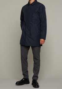 Abrigo impermeable azul marino con corte recto, que presenta un collar abotonado y bolsillos laterales, combinado con pantalones grises de estampado y zapatos negros.