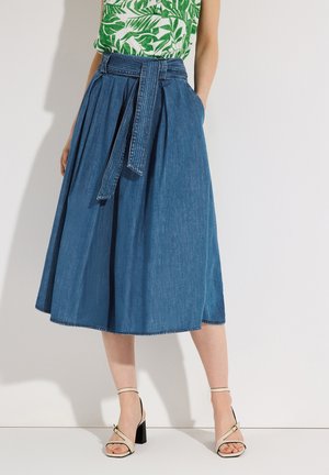 Frau trägt einen blauem Jeansrock mittlerer Länge mit Gürtel, ein grünes und weißes Oberteil mit Blattmuster und beigen Sandalen mit hohen Absätzen, stehend.