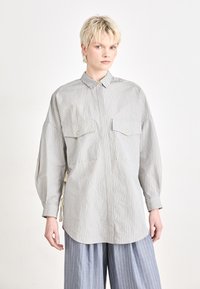 Camicia a righe con bottoni in grigio chiaro e crema, dotata di due tasche sul petto, colletto classico e maniche larghe. Righe verticali; texture morbida.