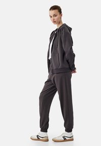 Mørkegrå hoodie og matchende joggers i blødt stof. Har en frontlomme, elastiske manchetter og hvide sneakers med sorte accenter.
