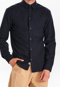 Granatowa koszula zapinana na guziki z teksturowanej tkaniny, z klasycznym kołnierzykiem, przednią kieszenią i kontrastowymi brązowymi guzikami. Noszona z beżowymi spodniami.