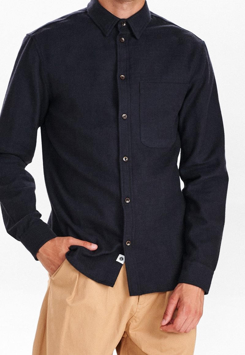 Granatowa koszula zapinana na guziki z teksturowanej tkaniny, z klasycznym kołnierzykiem, przednią kieszenią i kontrastowymi brązowymi guzikami. Noszona z beżowymi spodniami.