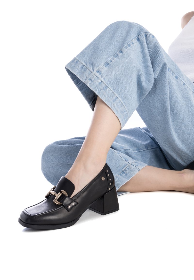 Mocassini con tacco in pelle nera, punta quadrata, ampia fascia e dettaglio con fibbia, con accenti metallici sul tacco. Indossati con pantaloni in denim blu chiaro.