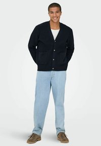 Marineblauer gerippter Cardigan mit V-Ausschnitt, Knopfverschluss und zwei Fronttaschen, kombiniert mit hellblauen Jeans und braunen Schuhen.