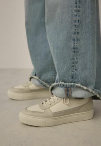 Weiße Leder-Sneaker mit strukturiertem Obermaterial, grauen Akzenten und einer klobigen Sohle, getragen mit hochgekrempelten hellblauen Jeans.