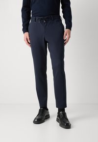 Άνδρας ντυμένος με μπλε σιδερωμένα παντελόνια με κορδόνι, μαύρες κοσμηματοποιημένες παπούτσια και ένα σκούρο μακρυμάνικο μπλουζάκι, στέκεται πάνω σε άσπρο δάπεδο και γκρι φόντο.