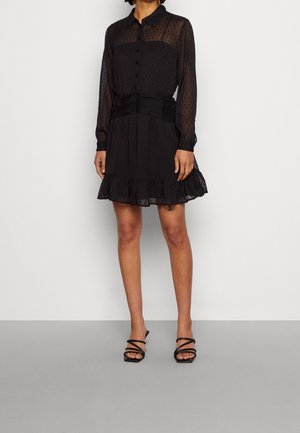 Kobieta ubrana w przezroczystą czarną sukienkę z długim rękawem, falbanami na dole oraz czarne sandały na ramiączkach, stojąca na tle gładkiej, białej ściany.