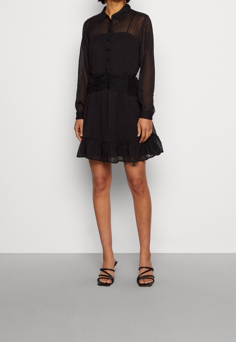 Femme portant une robe noire transparente à manches longues avec un ourlet à volants et des sandales noires à lanières, debout devant un fond blanc uni.