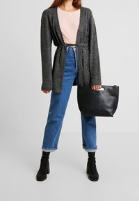Person som bär en grå bälte cardigan, rosa skjorta, uppvikta blå jeans, svarta klackstövlar och håller en svart läderhandväska.