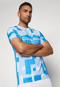 Svijetloplava sportska majica s apstraktnim uzorcima u tamnijim plavim tonovima. Ima kratke rukave i okrugli izrez. Logo i tekst na prednjoj strani.