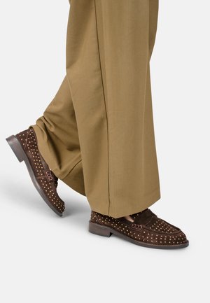 Persoon die wijde bruine broek en donkerbruine loafers met studs draagt, staand met één voet iets opgetild.