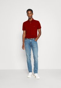 Polo shirt en coton rouge avec col, associé à un jean bleu clair. L'ensemble comprend une ceinture noire et des baskets blanches.