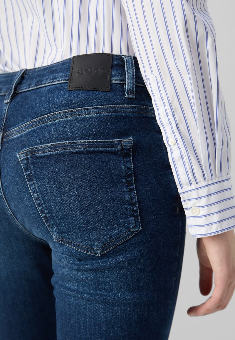 Jeans de mezclilla azul oscuro con un diseño ajustado, que presentan una etiqueta de cuero de BOSS en el bolsillo trasero y detalles de costura visibles. Camisa a rayas visible.