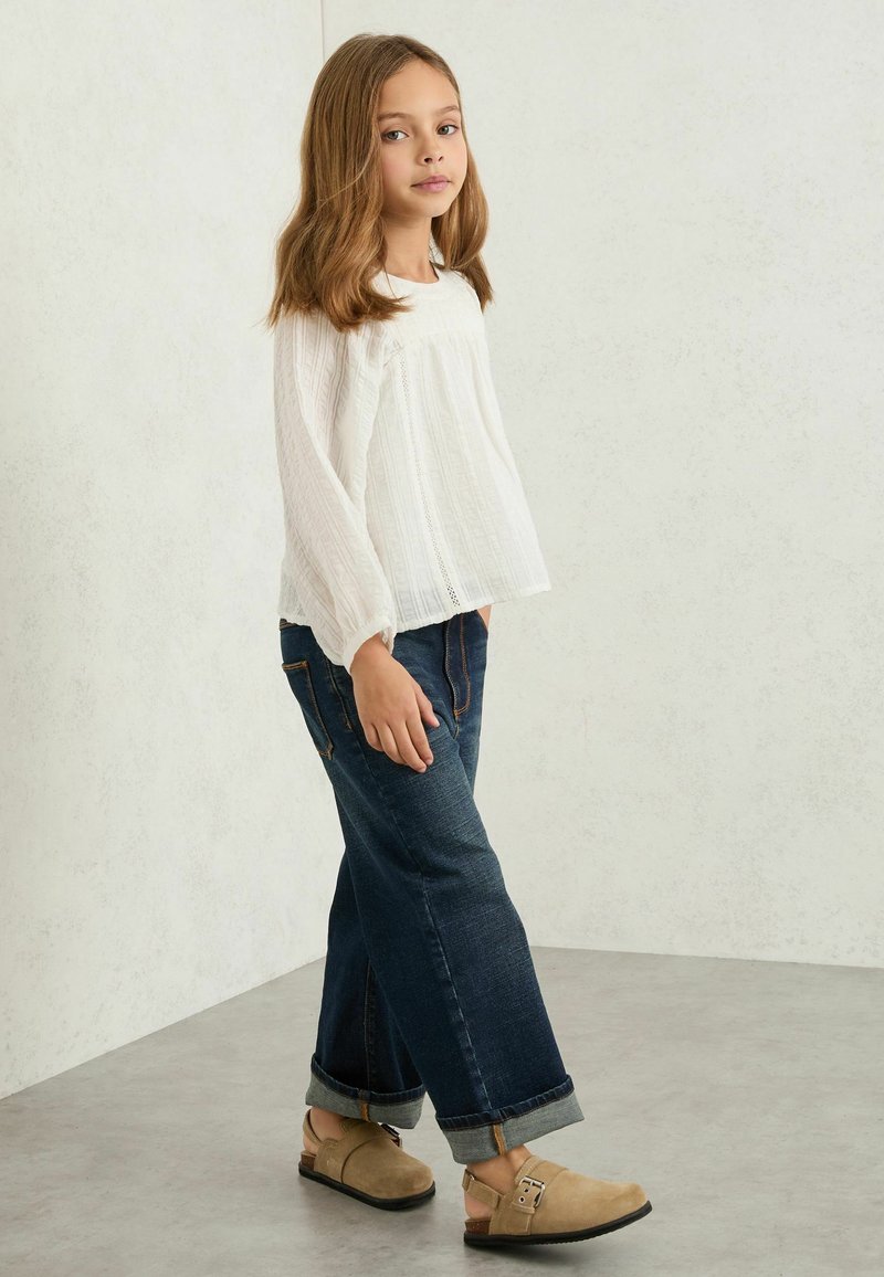 Junges Mädchen mit langen Haaren, das eine weiße Langarmbluse, hochgekrempelte dunkelblaue Jeans und beige Slip-on-Schuhe trägt, steht drinnen.