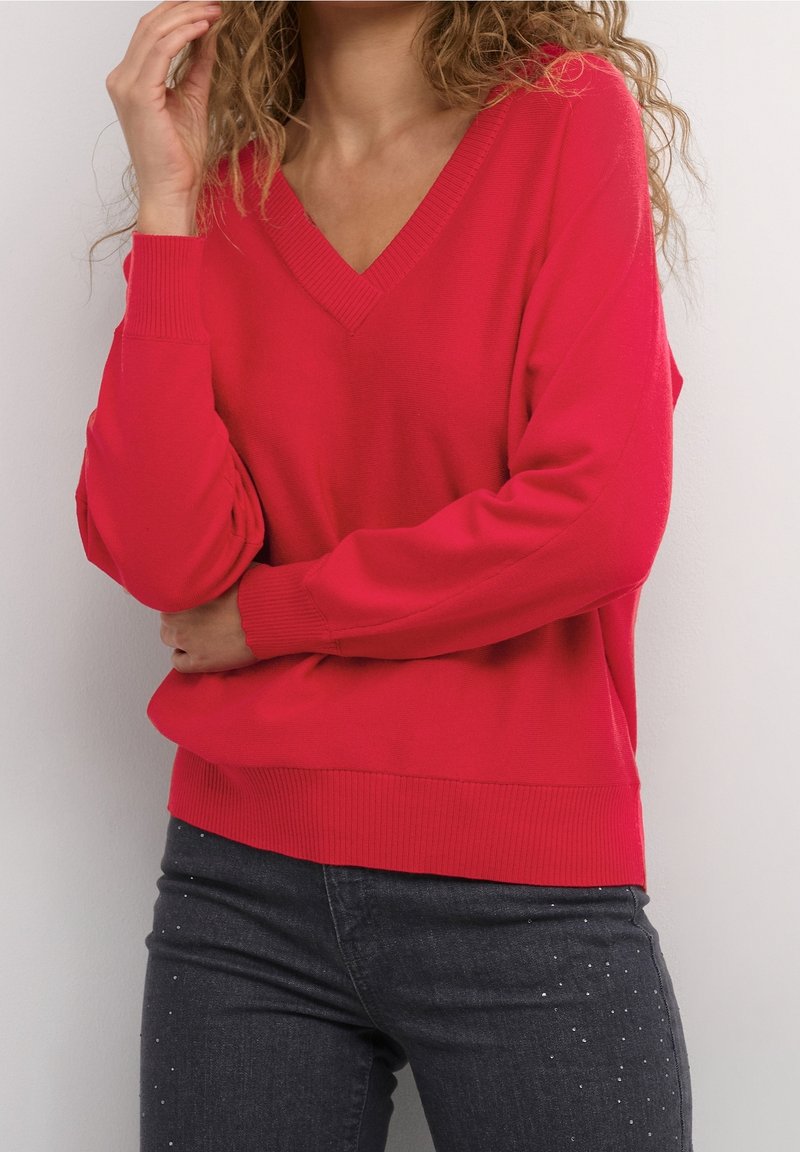 Eine Frau trägt einen knallrot V-Ausschnitt-Pullover und dunkle Jeans mit kleinen dekorativen Nieten. Sie posiert mit einer Hand in der Nähe ihres Gesichts und dem anderen Arm verschränkt.