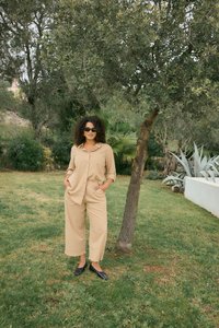 Kobieta o kręconych włosach i w okularach przeciwsłonecznych, ubrana w beżową koszulę i spodnie, stoi na trawie obok drzewa z zielonymi liśćmi w ogrodzie.