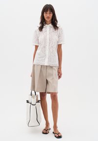 Blusa bianca a maniche corte con design ad occhio di pernice e orlo scallop, abbinata a pantaloni corti beige plissettati e sandali neri; con una borsa tote bianca.