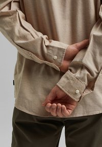 Personne portant une chemise beige à manches longues avec boutons, les mains jointes dans le dos, et un pantalon sombre sur un fond uni.