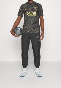 Maglia nera con design geometrico giallo, logo "QATAR AIRWAYS", abbinata a pantaloni neri Nike e scarpe da ginnastica multicolori, con in mano un pallone da calcio.