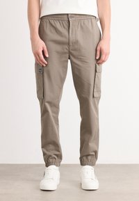 Spodnie cargo w kolorze khaki wykonane z bawełny, z elastycznym paskiem w talii, zwężającymi się mankietami na kostkach i kieszeniami bocznymi. Styl posiada naszywkę z logo.