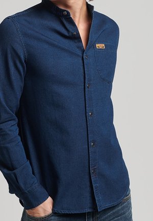 Homme portant une chemise en jean bleu foncé boutonnée avec une poche poitrine et une petite étiquette beige sur la poche, main dans la poche du jean.