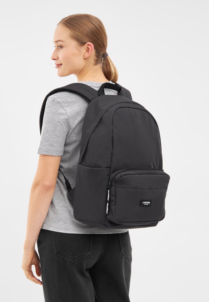 Black backpack with a durable fabric, rounded shape, front zip pocket, and adjustable shoulder straps. Minimalist design and soft texture.