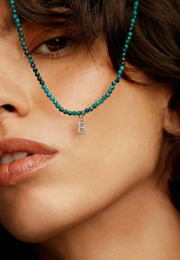 Collar de cuentas turquesa con un colgante de plata en forma de letra 'B', llevado sobre un tono de piel neutro, destacando las facciones del rostro.