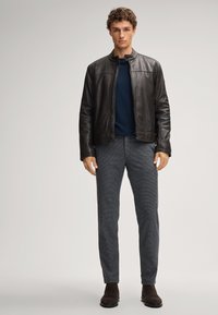 Mann steht in einer schwarzen Lederjacke, einem marineblauen Pullover, grauen karierten Hosen und dunkelorangen Wildlederstiefeln vor einem schlichten Hintergrund.