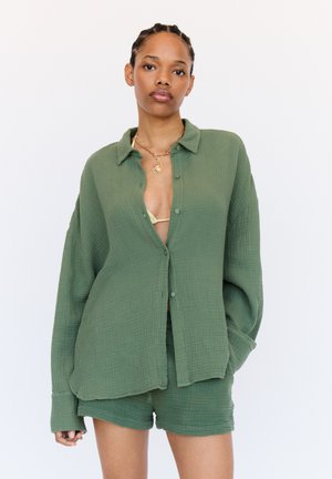 Olijfgroene, oversized, gestructureerde button-up shirt met een kraag, gecombineerd met een bijpassende short in een vergelijkbare stof en kleur.