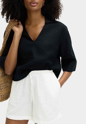 Kvinde iført en sort V-hals kortærmet top og hvide shorts med snøre, der holder en vævet brun skuldertaske med den ene hånd.